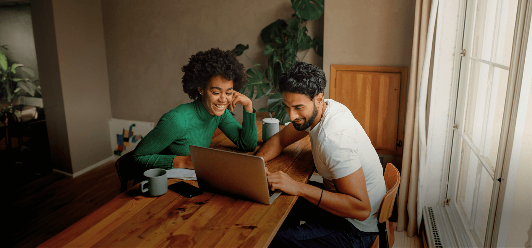 A smiling couple looking at a laptop together at a wooden dining table with coffee cups in a plant-filled apartment in Spain.