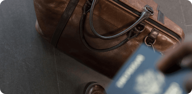 Close-up of a brown leather travel bag and a blurred passport in someone’s hand, suggesting travel readiness.