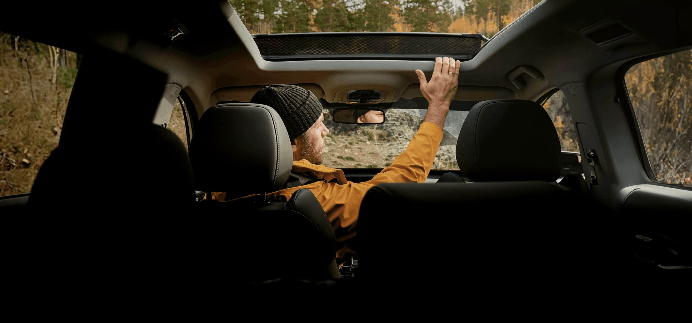 A man in a yellow jacket and beanie sitting in the driver's seat of a car, looking out at a forested mountain landscape.