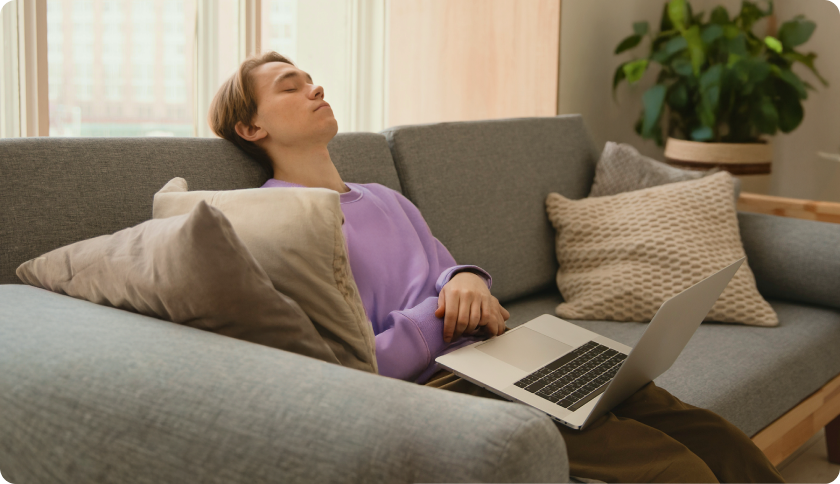 Person sitting on a couch with closed eyes, looking tired with a laptop on their knees.