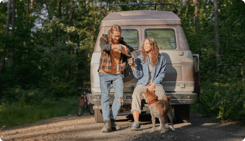 A couple with their dog sitting in front of their van in a forest, drinking coffee from a thermos.