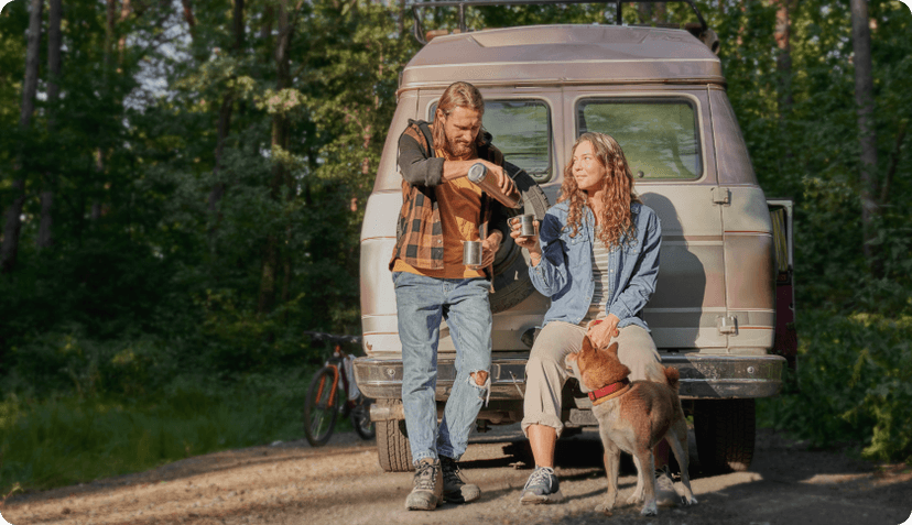 A couple with their dog sitting in front of their van in a forest, drinking coffee from a thermos.