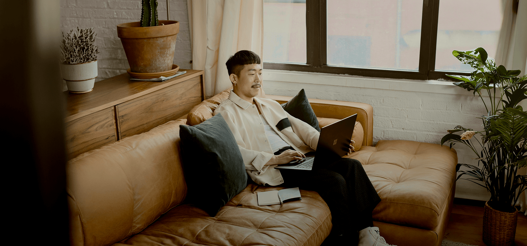A young man relaxing on a tan leather sofa with a laptop, surrounded by indoor plants in a sunlit apartment in France.