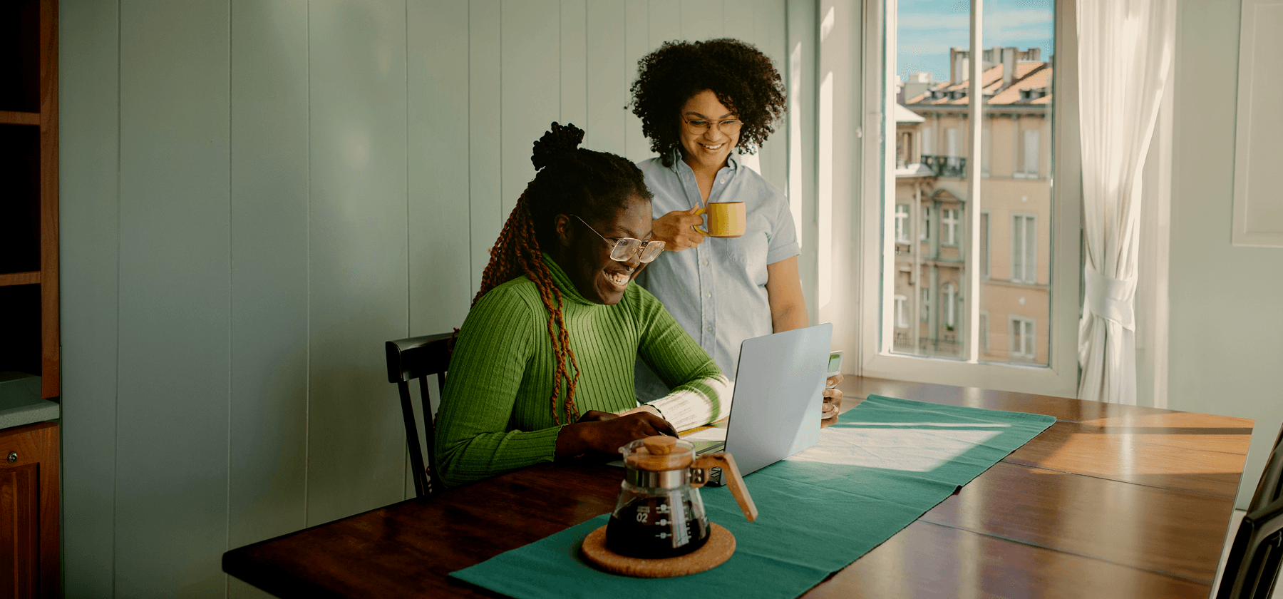 Two women smiling and looking at a laptop together at a dining table in a bright apartment in France.