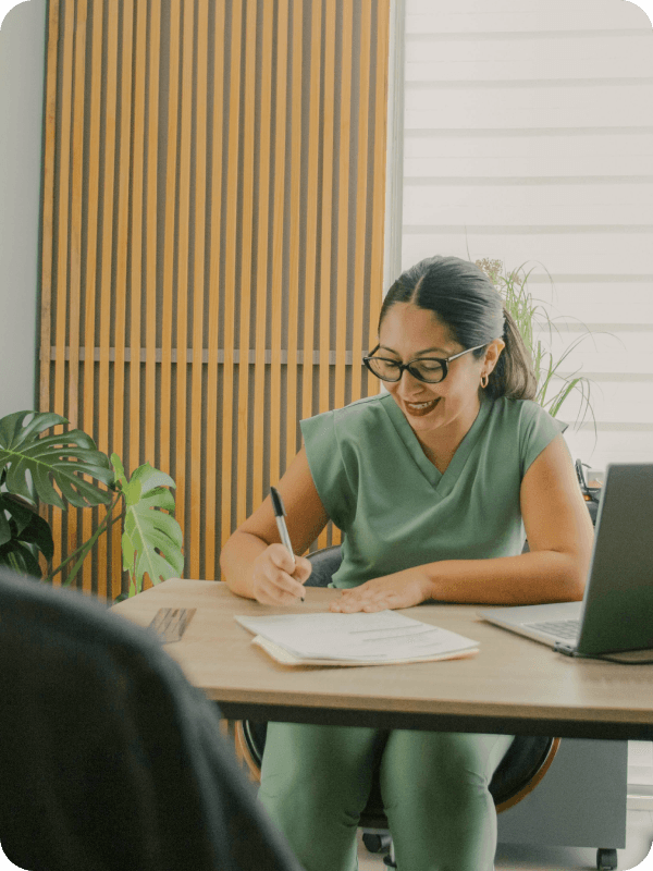 A doctor sitting at her office during a patient appointment, smiling and filling in documents.