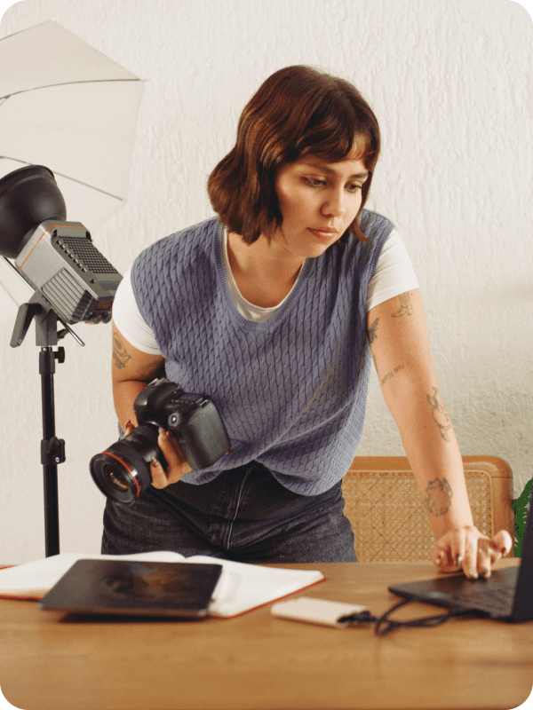 Photographer standing in front of her desk, holding a camera and checking her work on a laptop. Photoshoot equipment is seen in the background of the image.