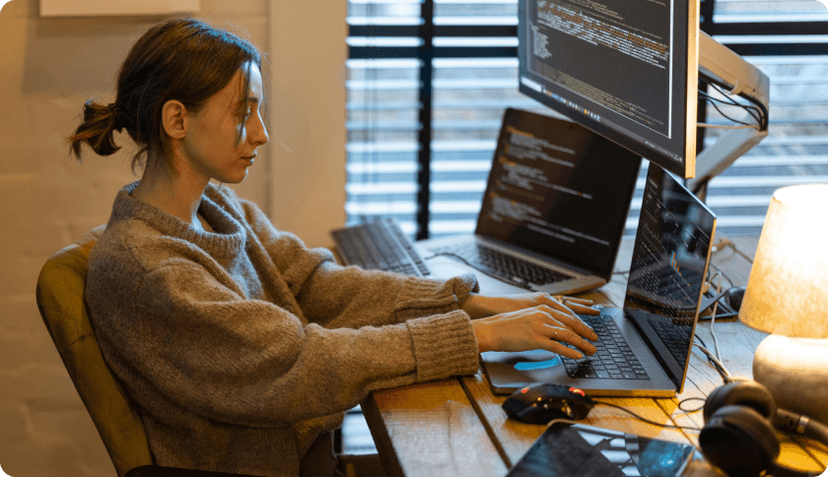 An IT freelancer working at her desk in front of 2 laptops and a screen showing lines of code.
