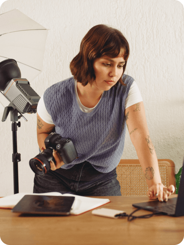 Photographer standing in front of her desk, holding a camera and checking her work on a laptop. Photoshoot equipment is seen in the background of the image.