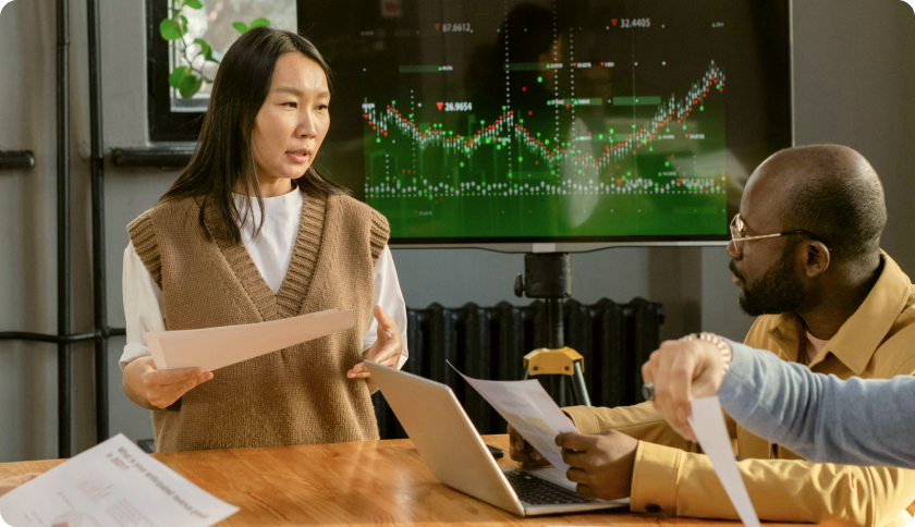A consultant talking to her client during a business meeting. The tv screen behind her shows graphs.