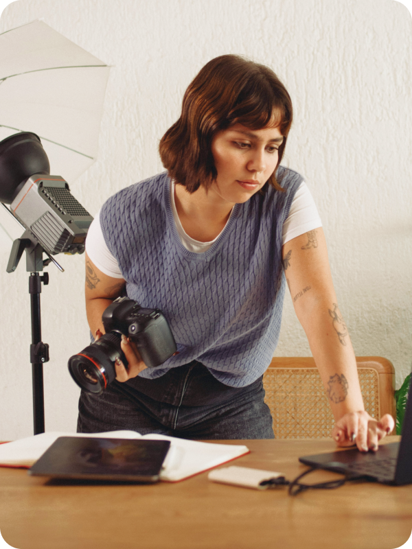 Photographer standing in front of her desk, holding a camera and checking her work on a laptop. Photoshoot equipment is seen in the background of the image.