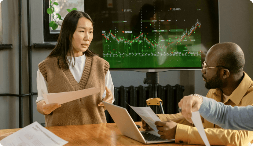 A consultant talking to her client during a business meeting. The tv screen behind her shows graphs.