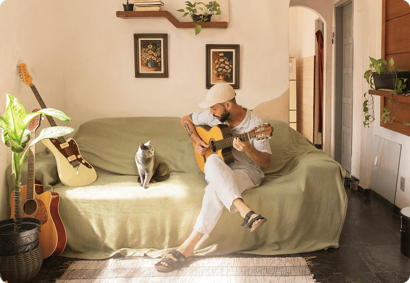 A man sitting on a couch in a cosy apartment, playing a guitar. A gray cat sits next to him.