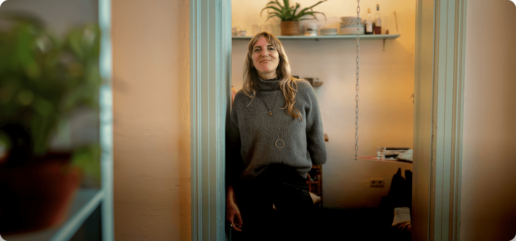 A woman smiling warmly while standing in the doorway of her cosy, plant-filled apartment.