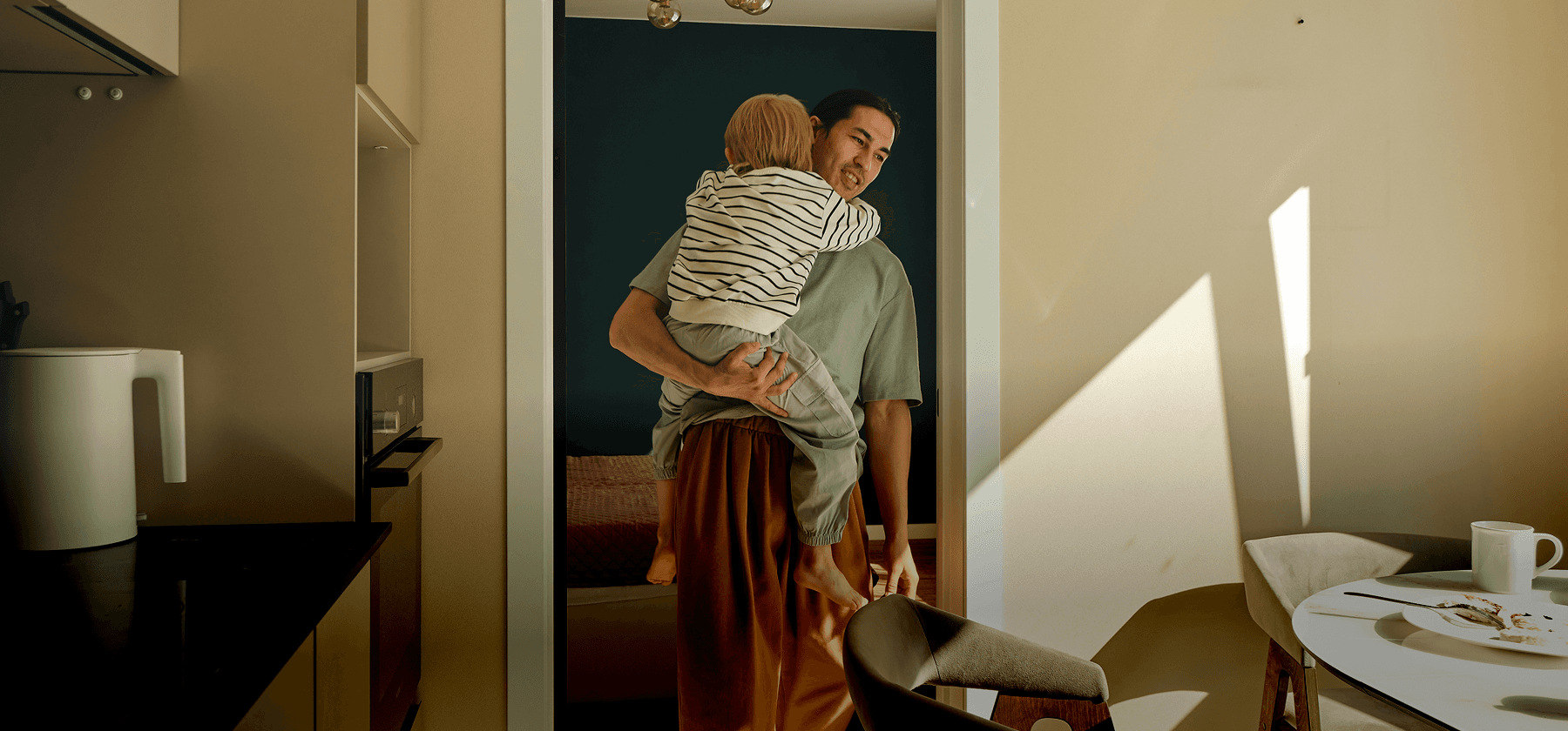 A father holding a young child in his arms in a sunlit modern apartment kitchen in France.