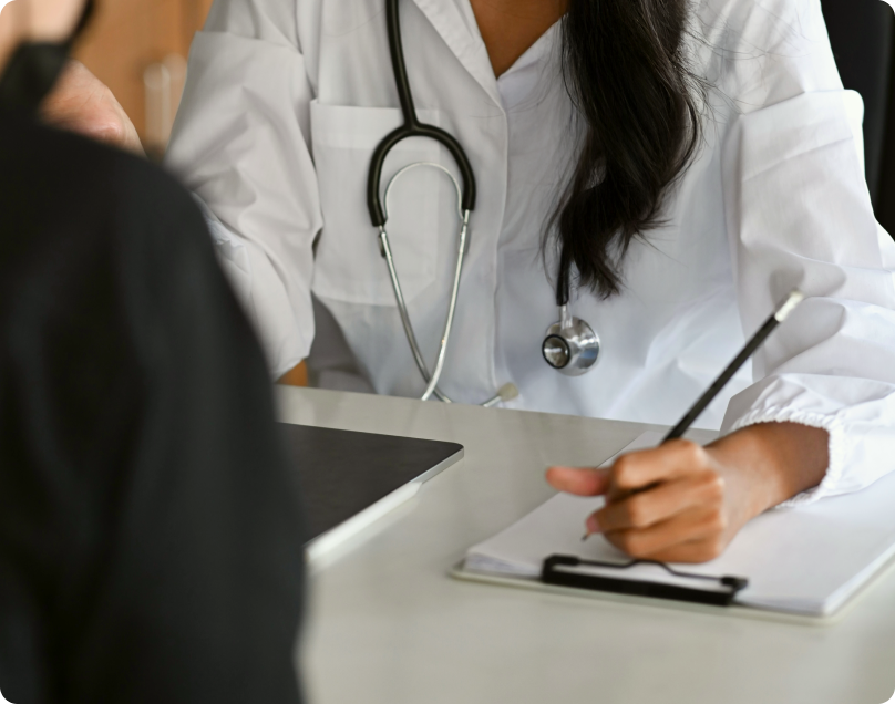 Doctor in scrubs with a stethoscope filling out a health questionnaire for a patient across the table.