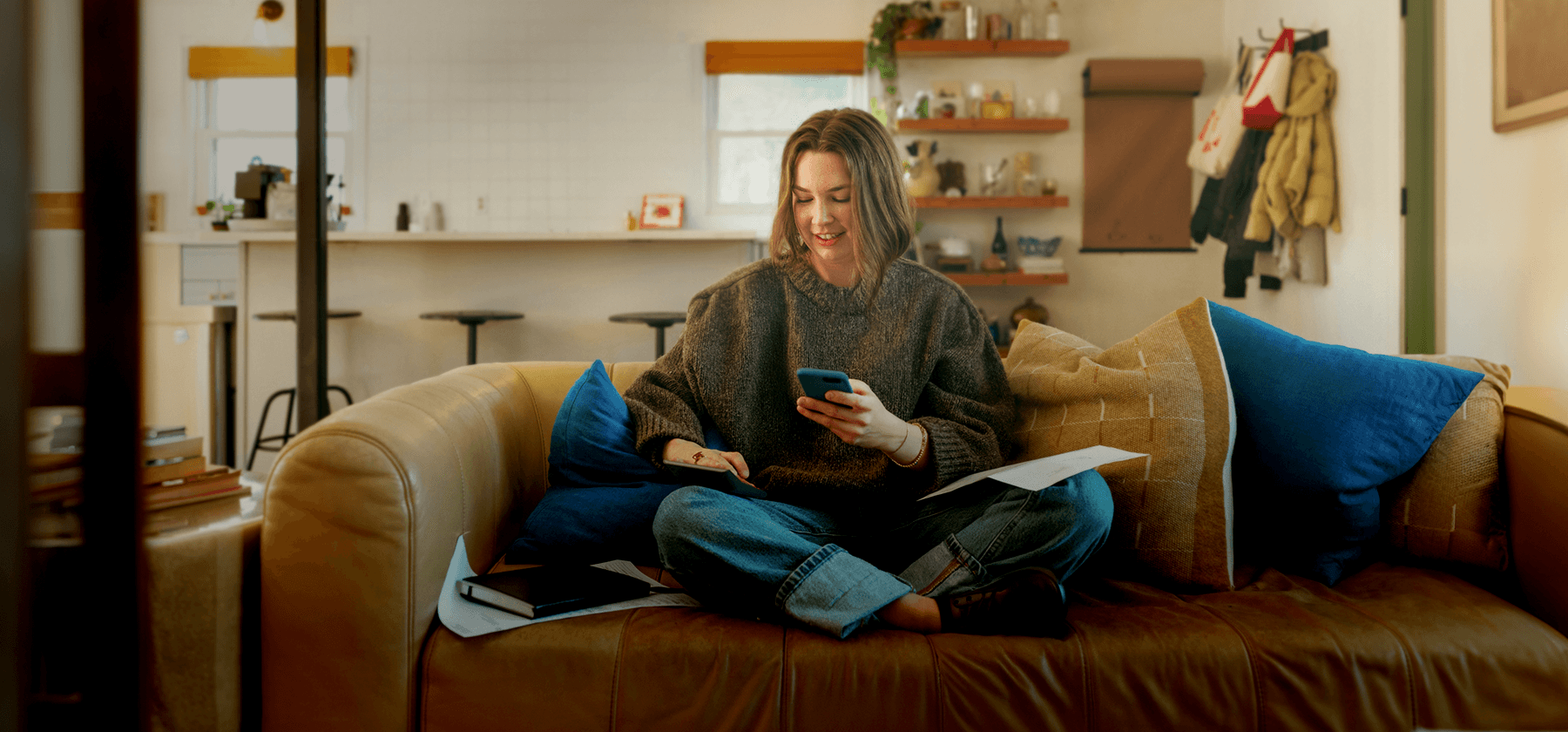 A woman sitting cross-legged on a sofa, smiling as she looks at her phone with papers in her lap.
